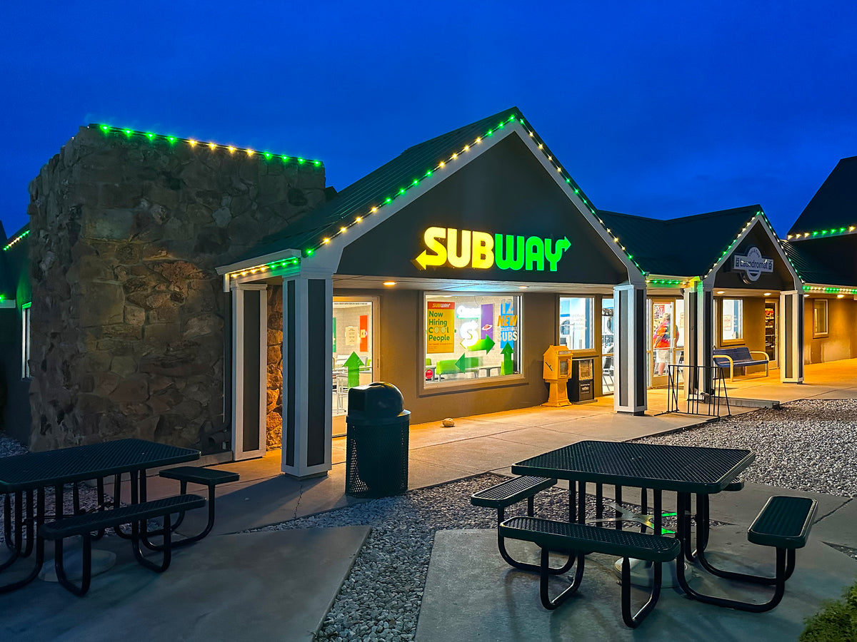 Green and warm white commercial roofline lighting installed on a Subway restaurant building at dusk.